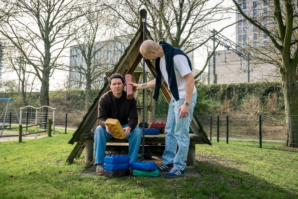 Jasper zit op een houten klimtoestel in een speeltuin en krijgt een bouwblok aangereikt van Pim, die ernaast staat